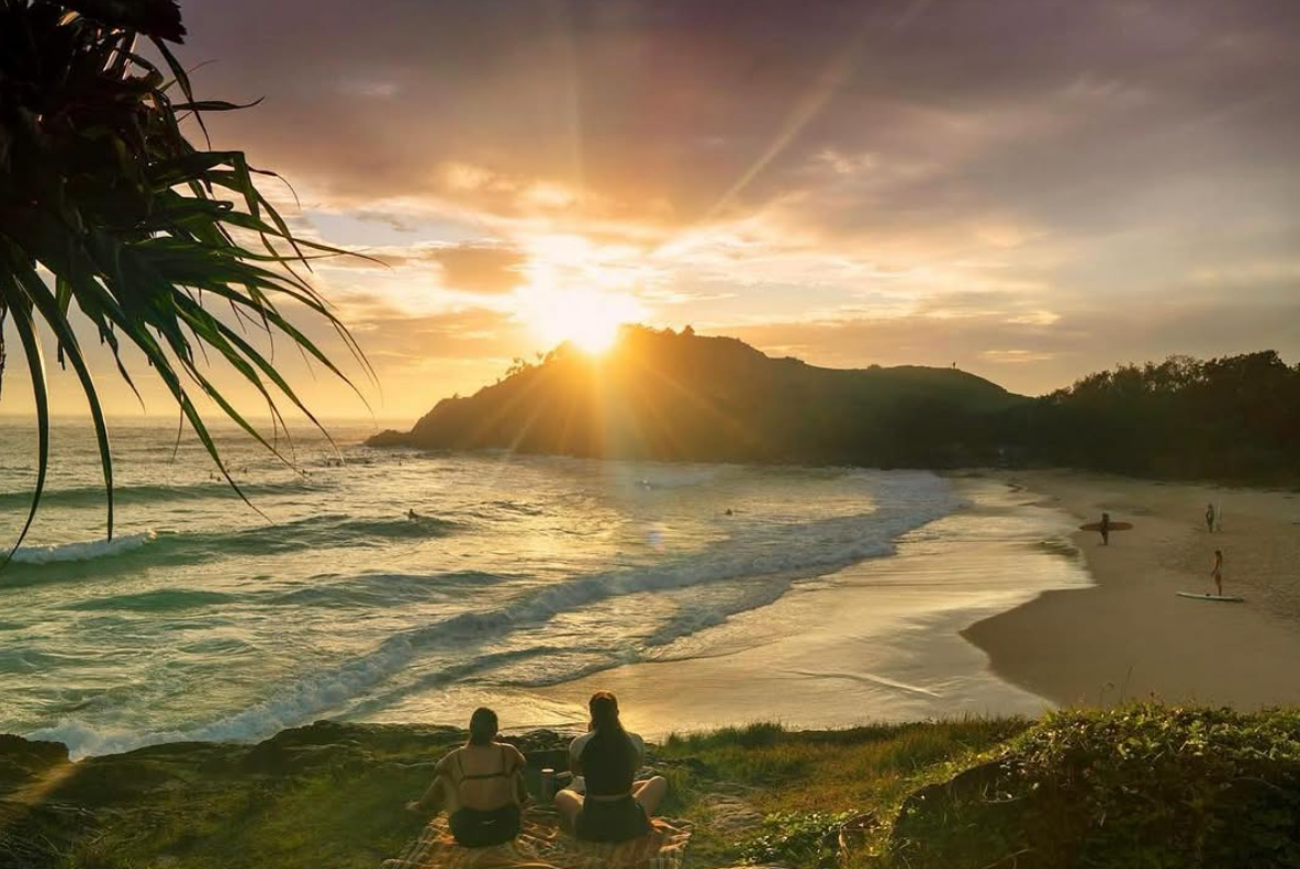 Sunset over a beach with people sitting on rocks and trees in the foreground.